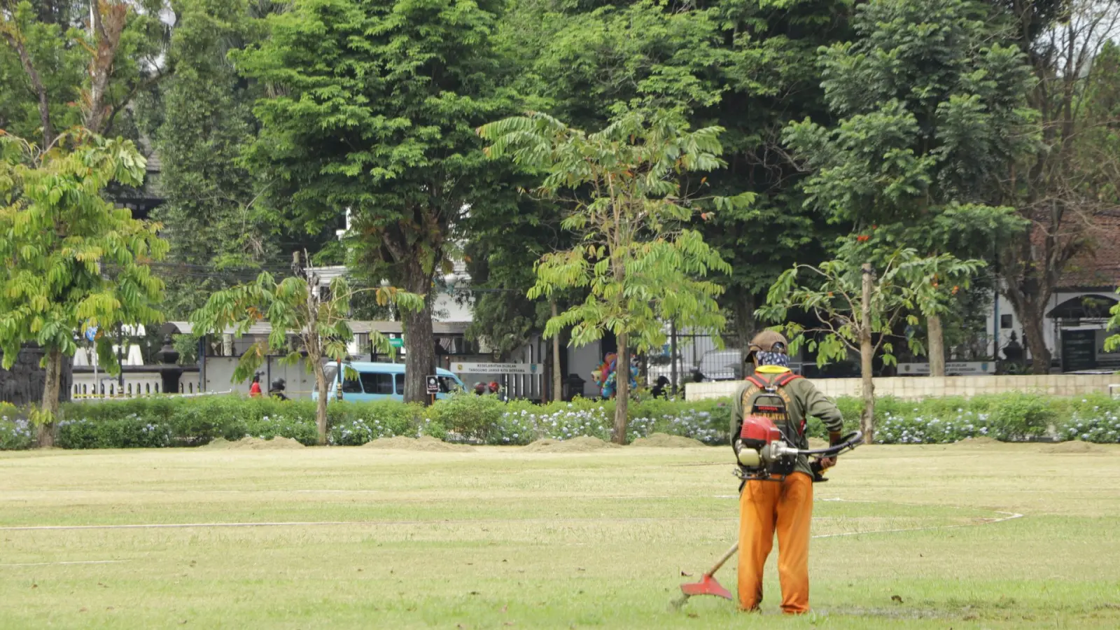 Groundskeeper trimming a large park lawn, seen from a distance, during scheduled commercial landscape maintenance.